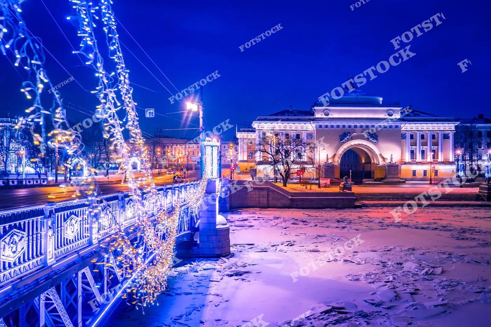 На фотографии изображен празднично украшенный мост через реку, увешанный гирляндами и огнями. Вдалеке видно здание Адмиралтейства, подсвеченное теплой иллюминацией. Река покрыта льдом, что подчеркивает зимний характер снимка. Ночная съемка добавляет снимку загадочности и волшебства, характерных для зимнего Петербурга.